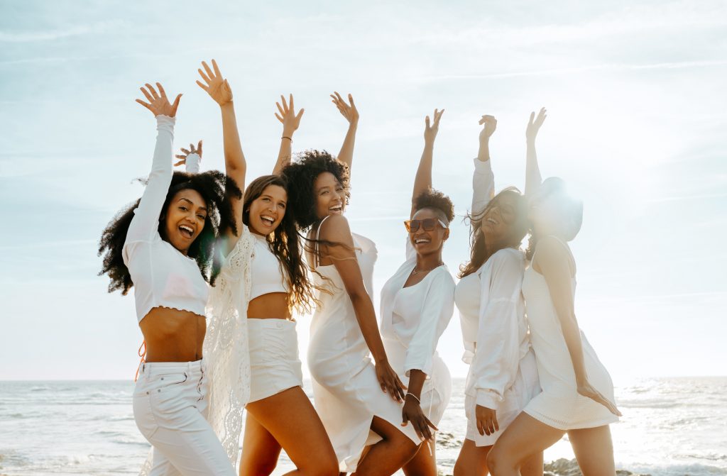 Group of happy female friends cheering with their arms raised while celebrating hen party at the beach smiling at camera