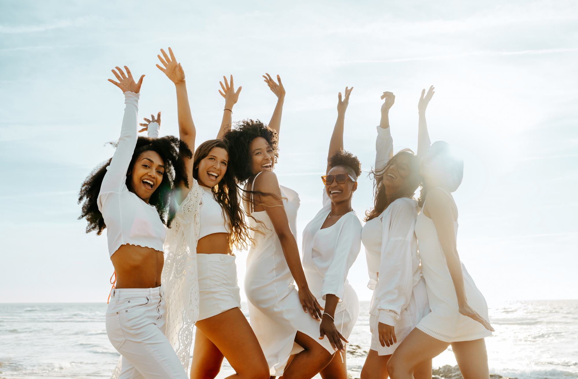 Group of happy female friends cheering with their arms raised while celebrating hen party at the beach smiling at camera